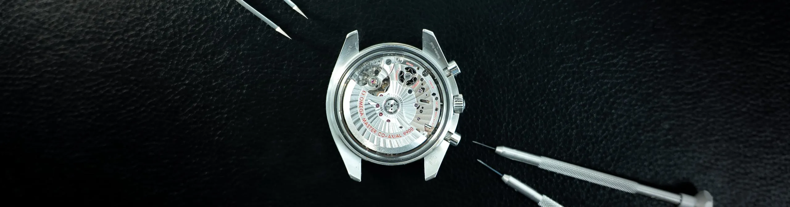 Omega Speedmaster caseback showing the Master Co-Axial 9900 movement, with screwdrivers on the SwissMade bench